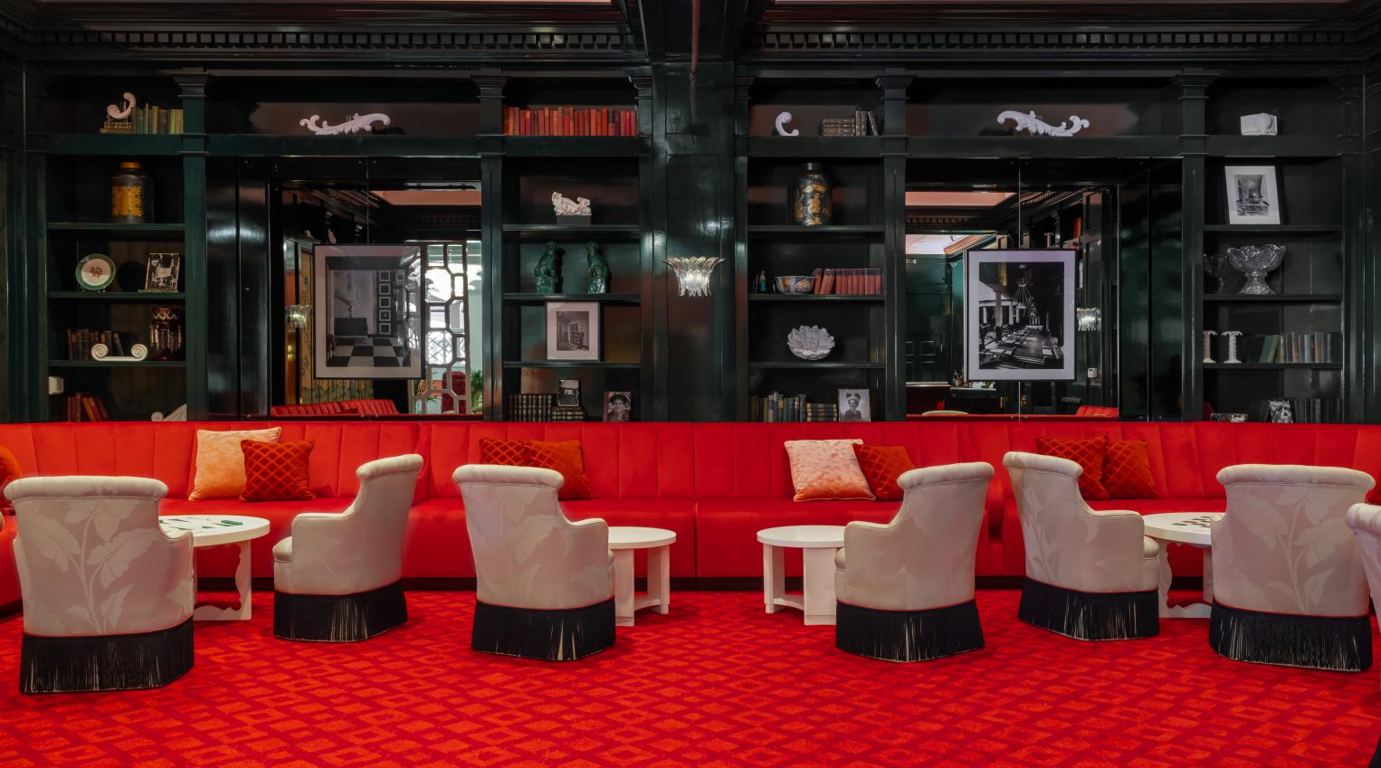 Luxurious lounge with red velvet seating, patterned carpet, cream armchairs, and black lacquered shelving in an elegant upscale interior.