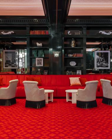Luxurious lounge with red velvet seating, patterned carpet, cream armchairs, and black lacquered shelving in an elegant upscale interior.