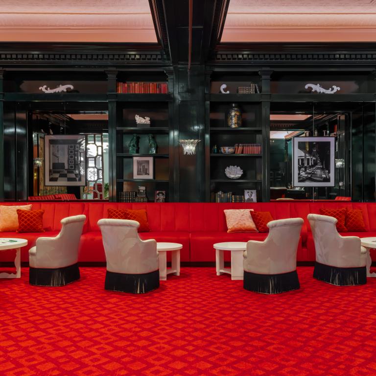 Luxurious lounge with red velvet seating, patterned carpet, cream armchairs, and black lacquered shelving in an elegant upscale interior.