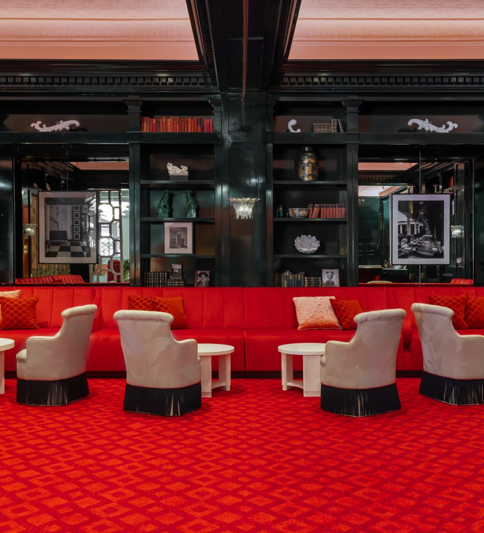 Luxurious lounge with red velvet seating, patterned carpet, cream armchairs, and black lacquered shelving in an elegant upscale interior.