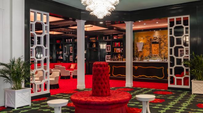 Luxurious lounge with red velvet seating, patterned carpet, cream armchairs, and black lacquered shelving in an elegant upscale interior.