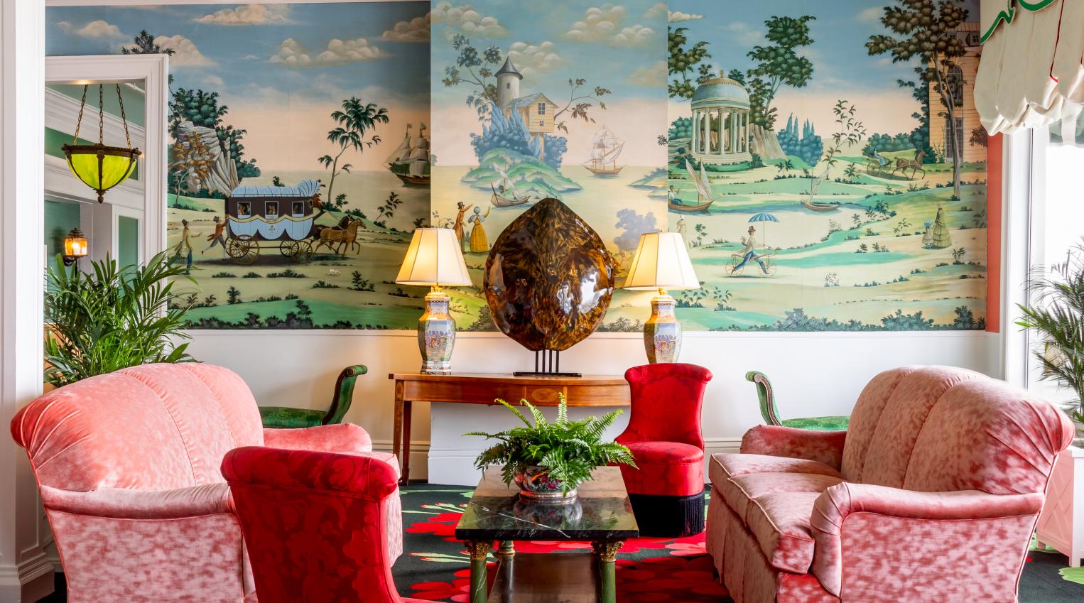 The vibrant and eclectic lobby of Grand Hotel, showcasing the iconic interior design by Carleton Varney, with its scenic mural wallpaper and bold pink and red furniture.