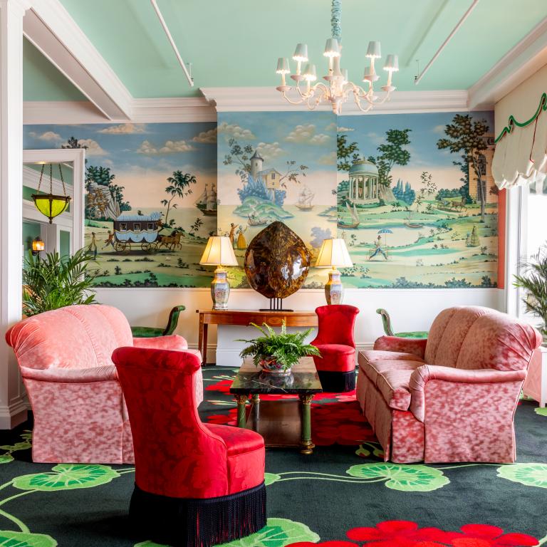 The vibrant and eclectic lobby of Grand Hotel, showcasing the iconic interior design by Carleton Varney, with its scenic mural wallpaper and bold pink and red furniture.