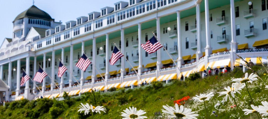 A beautiful low-angle view of the historic Grand Hotel on Mackinac Island, looking up through a field of white daisies on a sunny summer day.