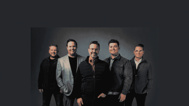 Six men in smart casual attire posing together for a group photo against a dark backdrop, smiling at the camera
