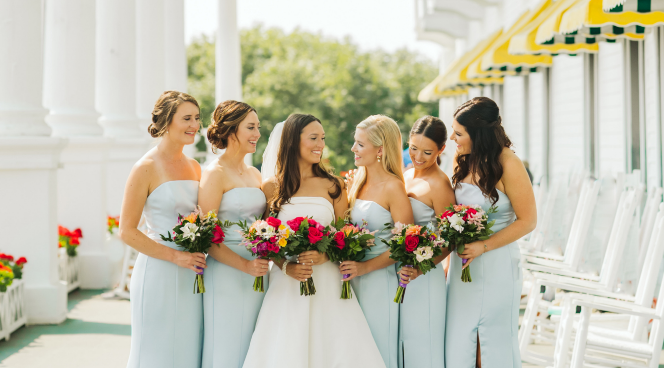 Group of bridesmaids in pale blue dresses holding colorful bouquets, standing together outdoors under white columns and yellow awnings.