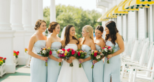 Group of bridesmaids in pale blue dresses holding colorful bouquets, standing together outdoors under white columns and yellow awnings.