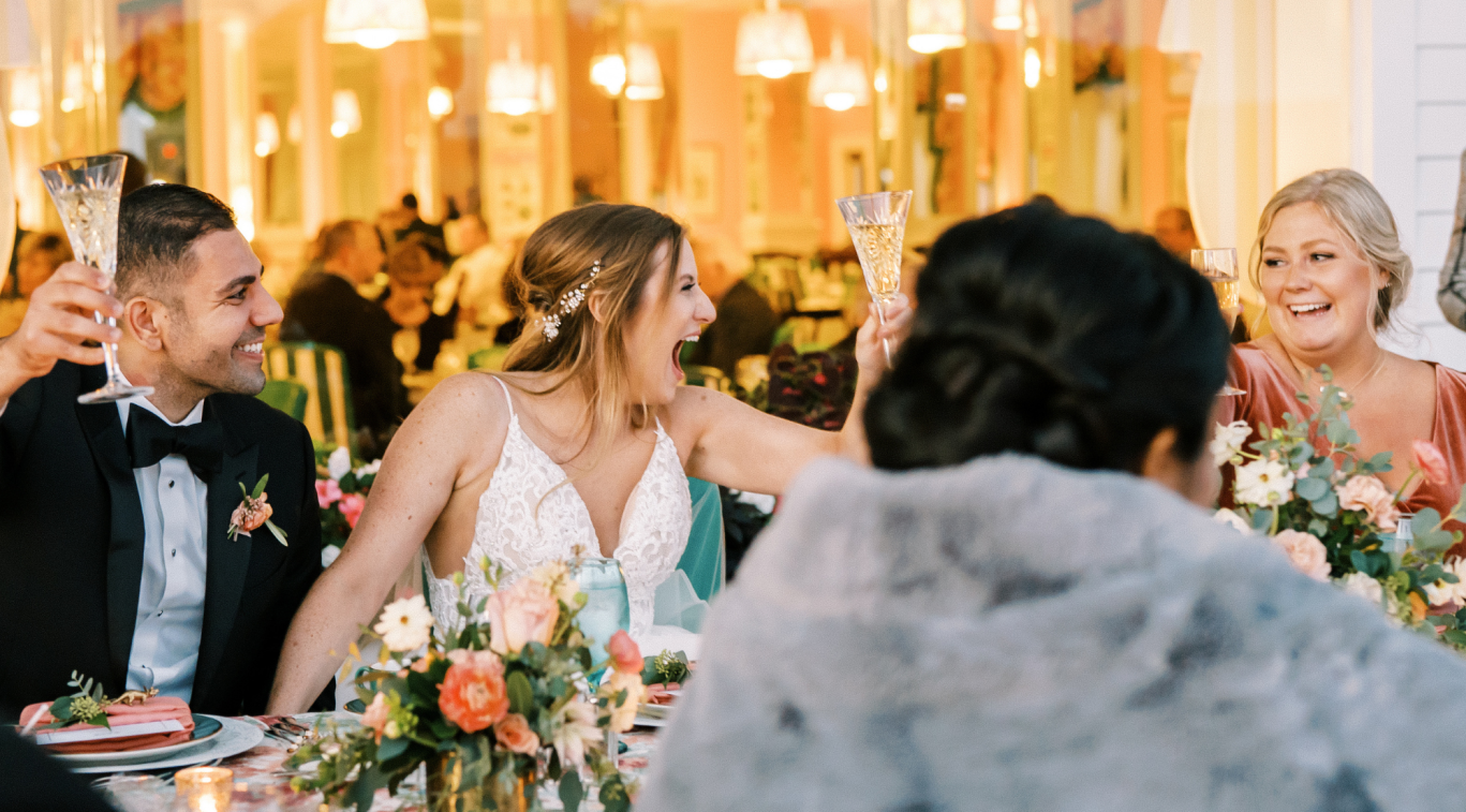 Bride in a lace wedding gown raises a glass with friends at a festively decorated reception table filled with flowers and candles, smiling and toasting.