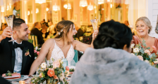Bride in a lace wedding gown raises a glass with friends at a festively decorated reception table filled with flowers and candles, smiling and toasting.