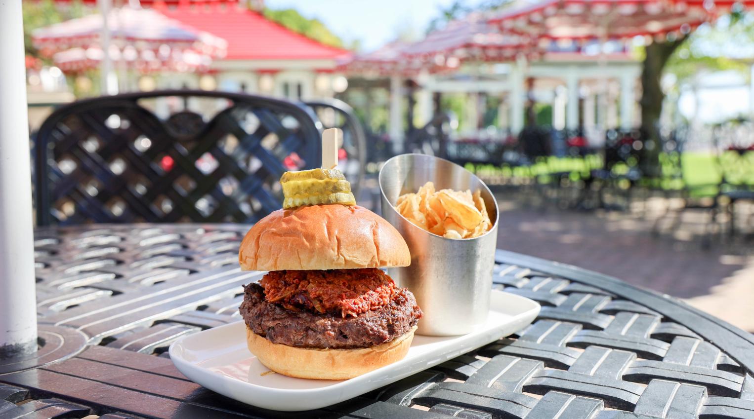 Two stacked burgers with a pickle spear on the top bun, served with fries in a metal cup on an outdoor patio.