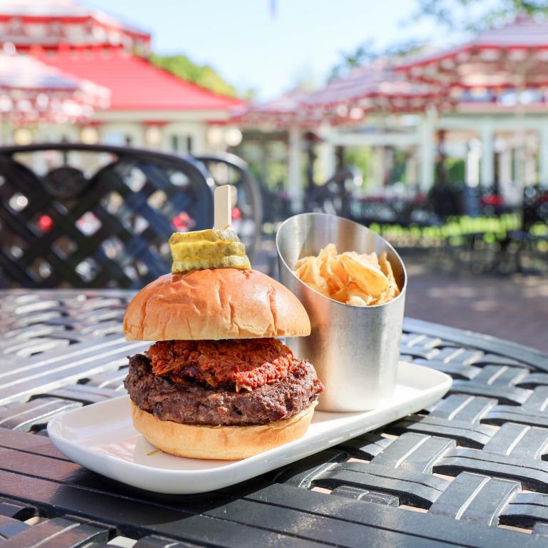 Two stacked burgers with a pickle spear on the top bun, served with fries in a metal cup on an outdoor patio.